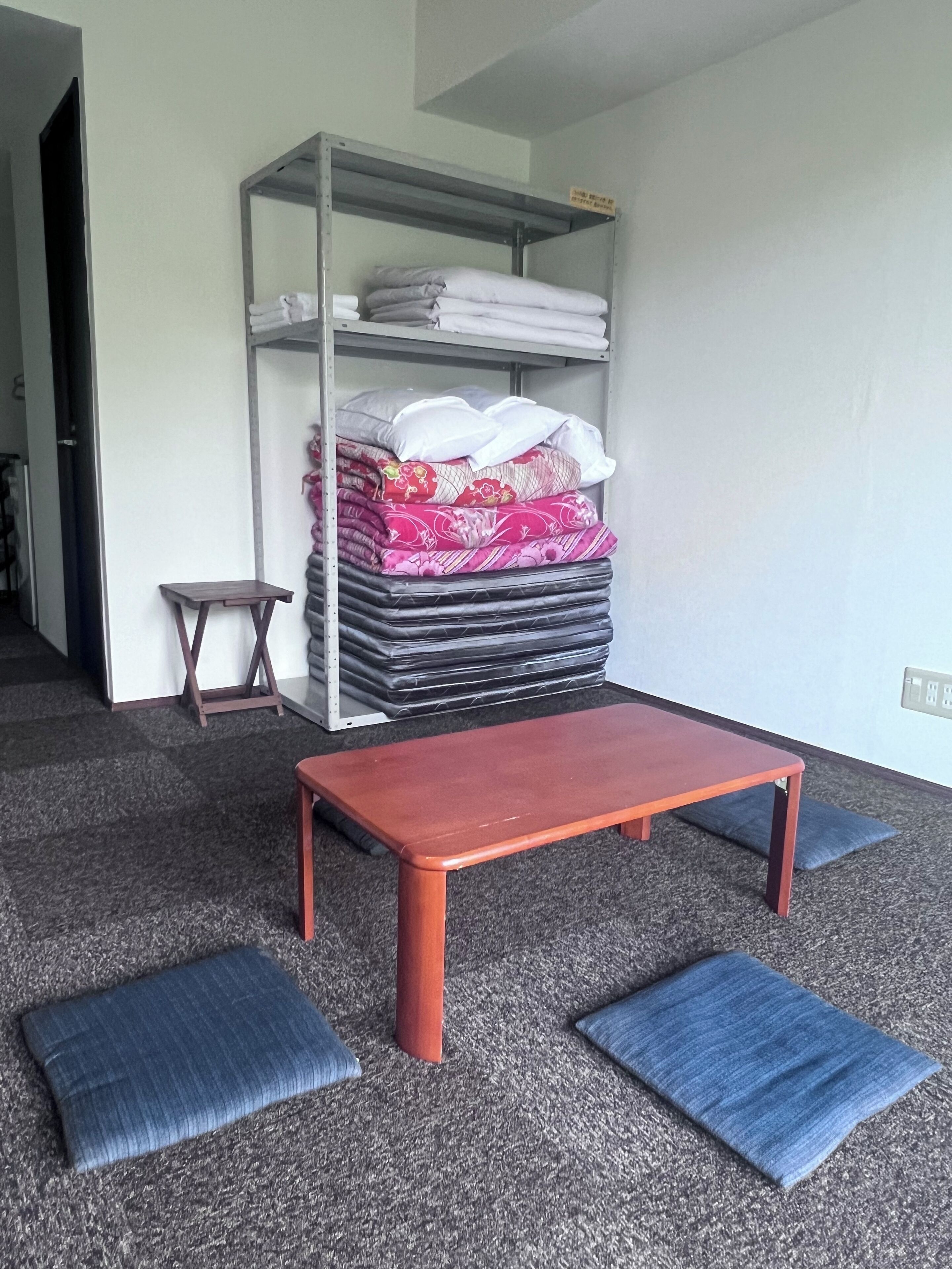 A room with a metal shelving unit storing folded bedding, a small wooden stool, a red bench, and two blue cushions on the carpeted floor.