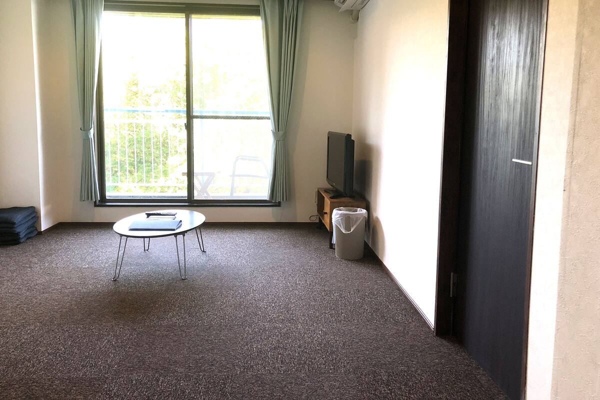 Ein Zimmer mit einem großen Fenster, einem kleinen runden Tisch, einem Fernseher auf einem Holzständer und einer Mülltonne.