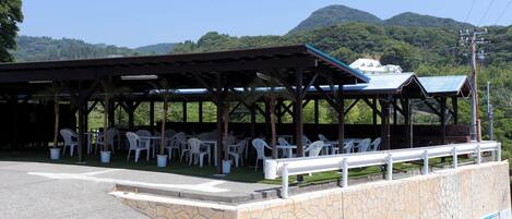 A covered outdoor seating area with white plastic chairs and tables, a stone wall, and a view of a mountain.