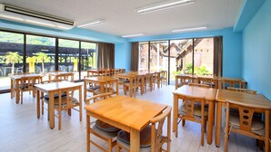 A spacious room with multiple wooden tables and chairs, large windows with a view of greenery and buildings, and a ceiling with fluorescent lighting. - Pension Bigfisher Club (Shimoda)