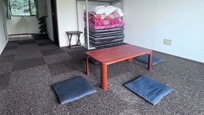 A room with a carpeted floor, a wooden table, two blue cushions, a metal shelf with folded blankets, and a window with curtains. - Pension Bigfisher Club (Shimoda)