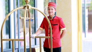 Un membre du personnel d’un hôtel, vêtu d’un uniforme rouge, se tient à côté d’un ascenseur doré et richement décoré.