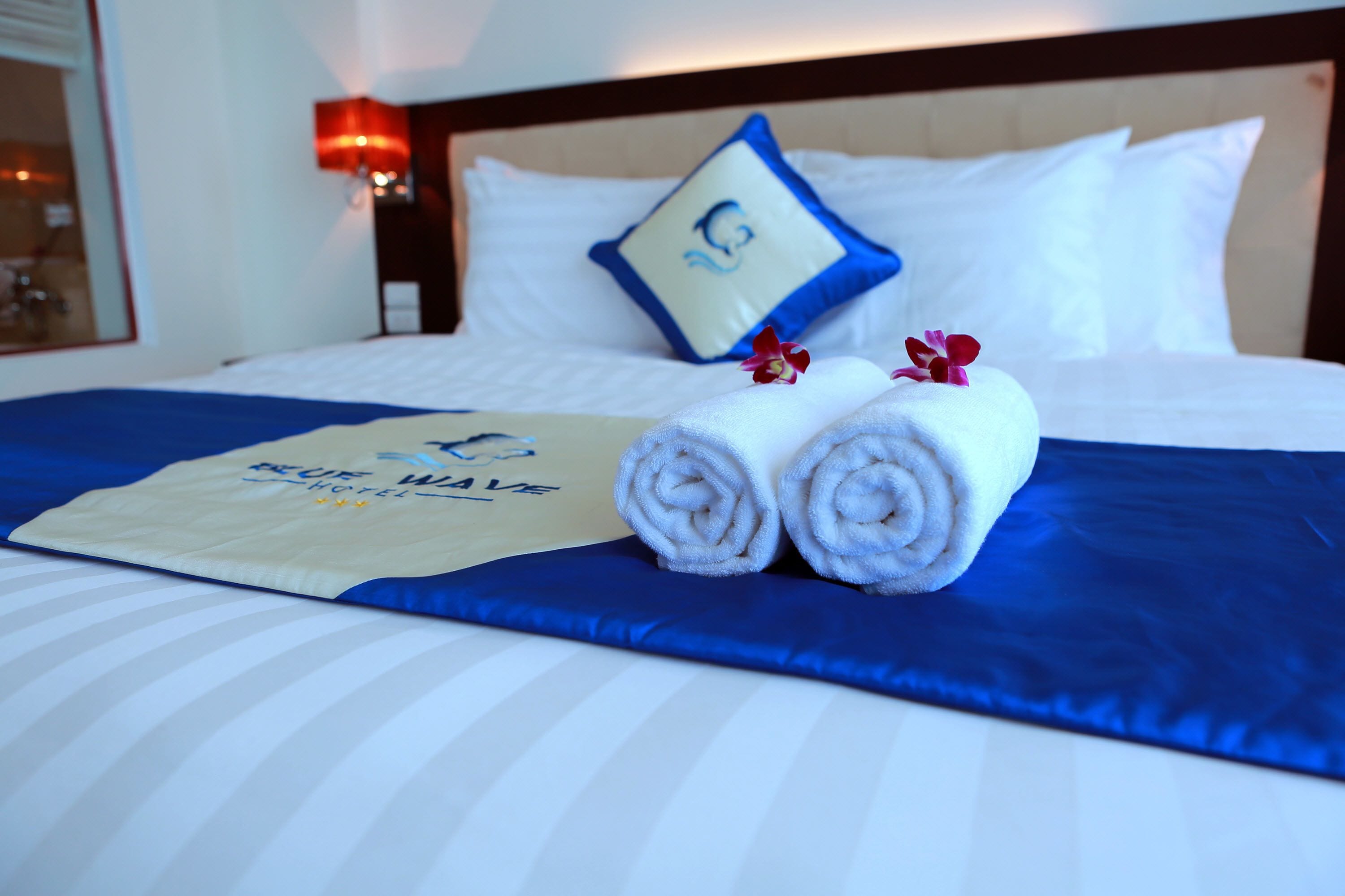 Una cama bien hecha con una colcha azul y blanca a rayas, una toalla doblada, un almohadón con un logotipo, y una habitación con una mesita de noche y una lámpara.
