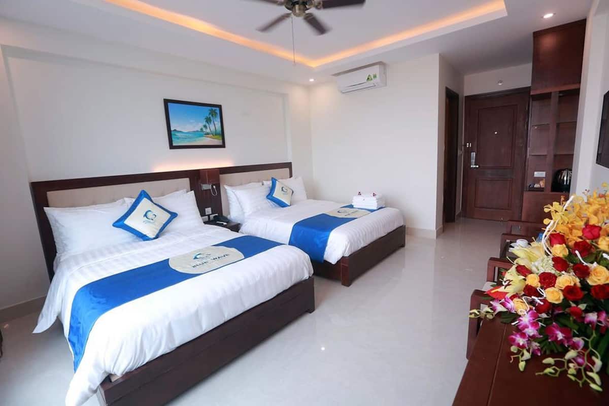 A hotel room with two beds, a ceiling fan, an air conditioning unit, a desk with a vase of flowers, and a door leading to a bathroom.