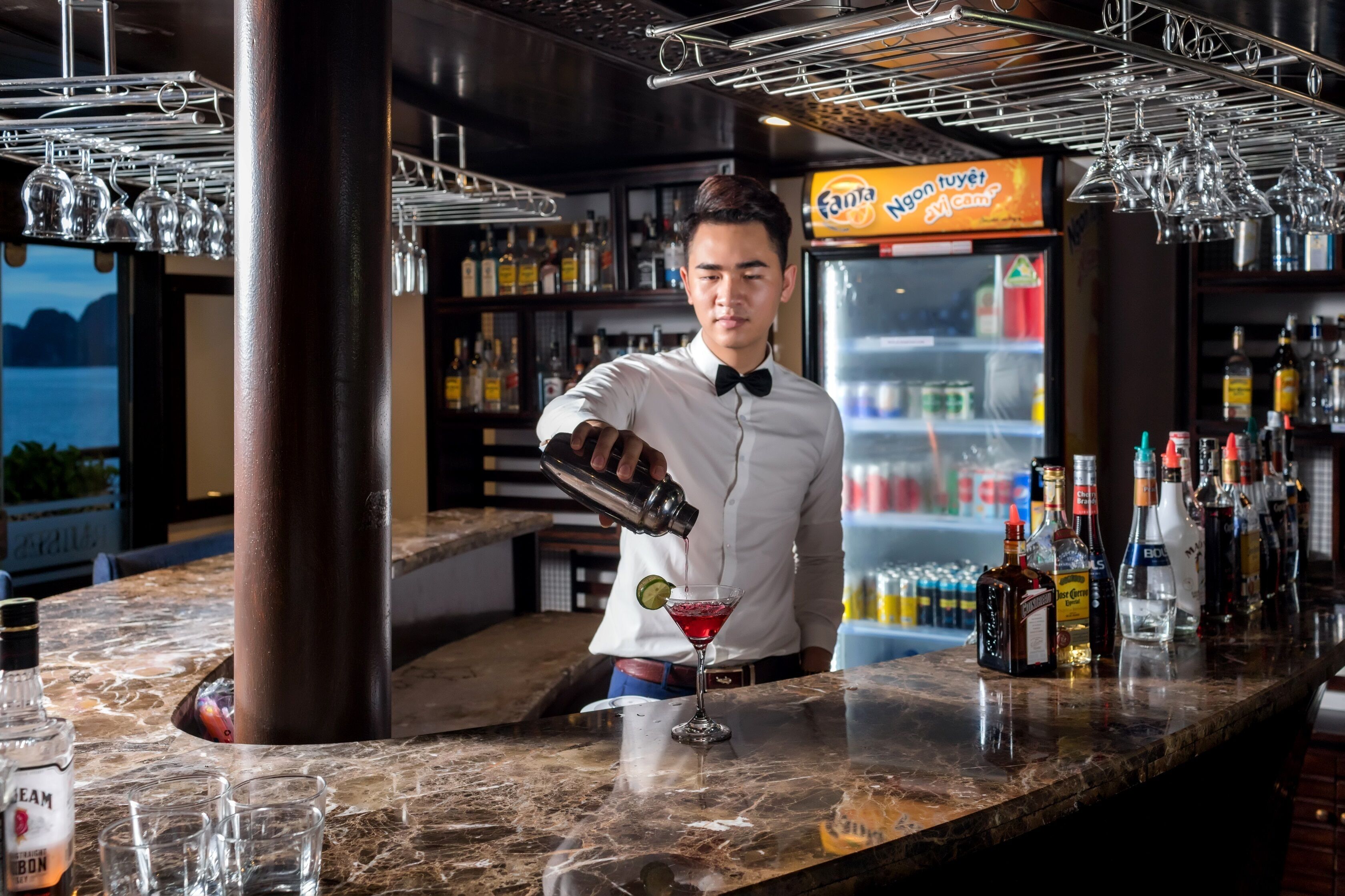 Un barman verse une boisson dans un verre à martini dans un bar doté d’un comptoir en marbre, d’étagères remplies de bouteilles et d’un réfrigérateur vitrine.