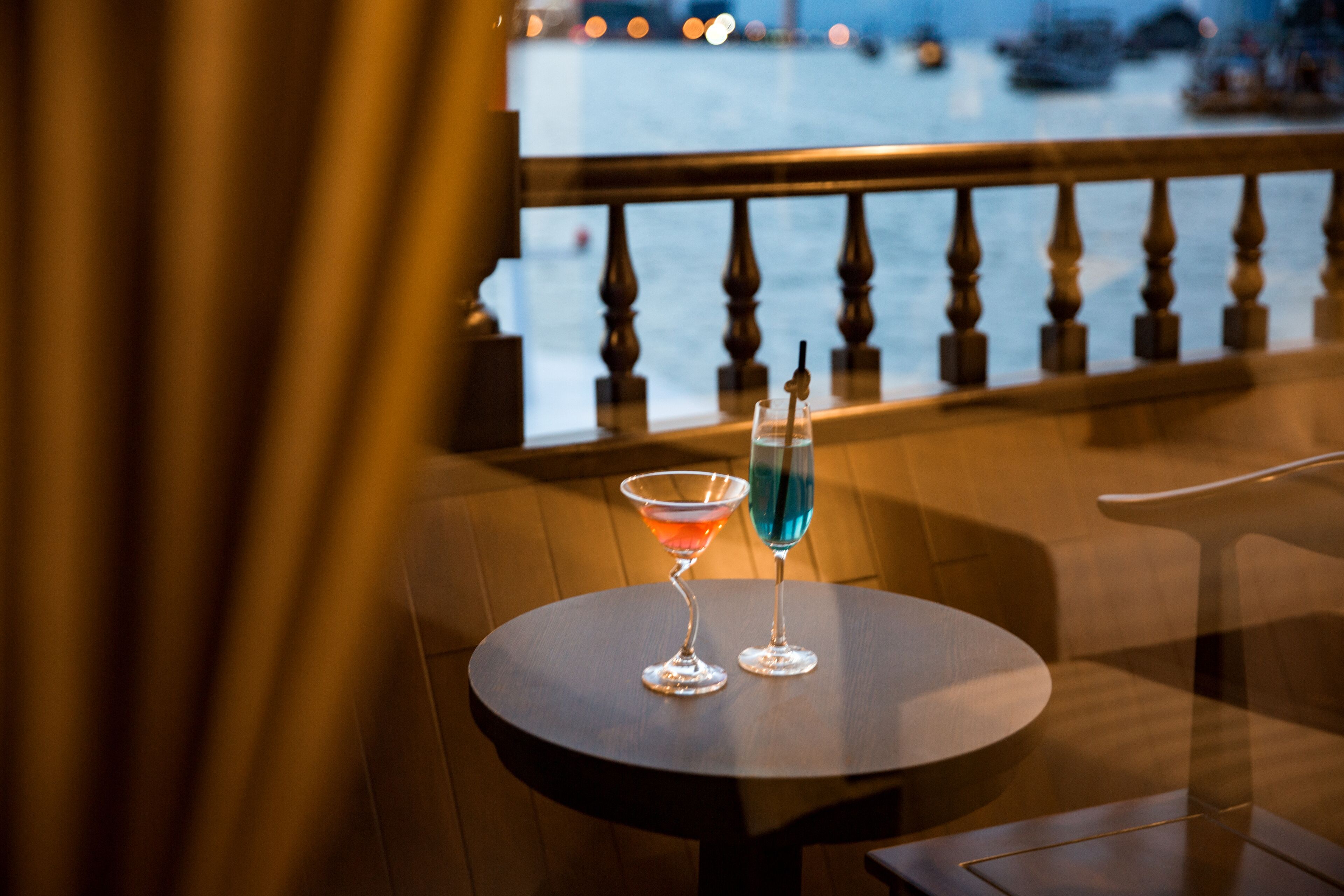 A round table with two glasses of cocktails on a balcony overlooking a body of water.