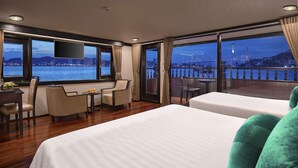 Une chambre d’hôtel dotée d’un grand lit, d’un bureau avec une chaise, d’une petite table avec deux chaises et d’un balcon offrant une vue sur l’eau et le panorama de la ville.