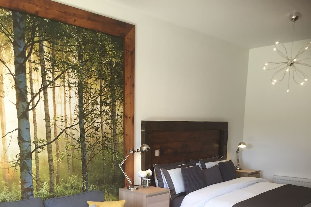 Une chambre avec un grand décor mural représentant une forêt, un lit avec une tête de lit sombre, une table de chevet avec des lampes, un canapé avec des coussins et une table basse en bois ornée d’un vase et d’un panier.