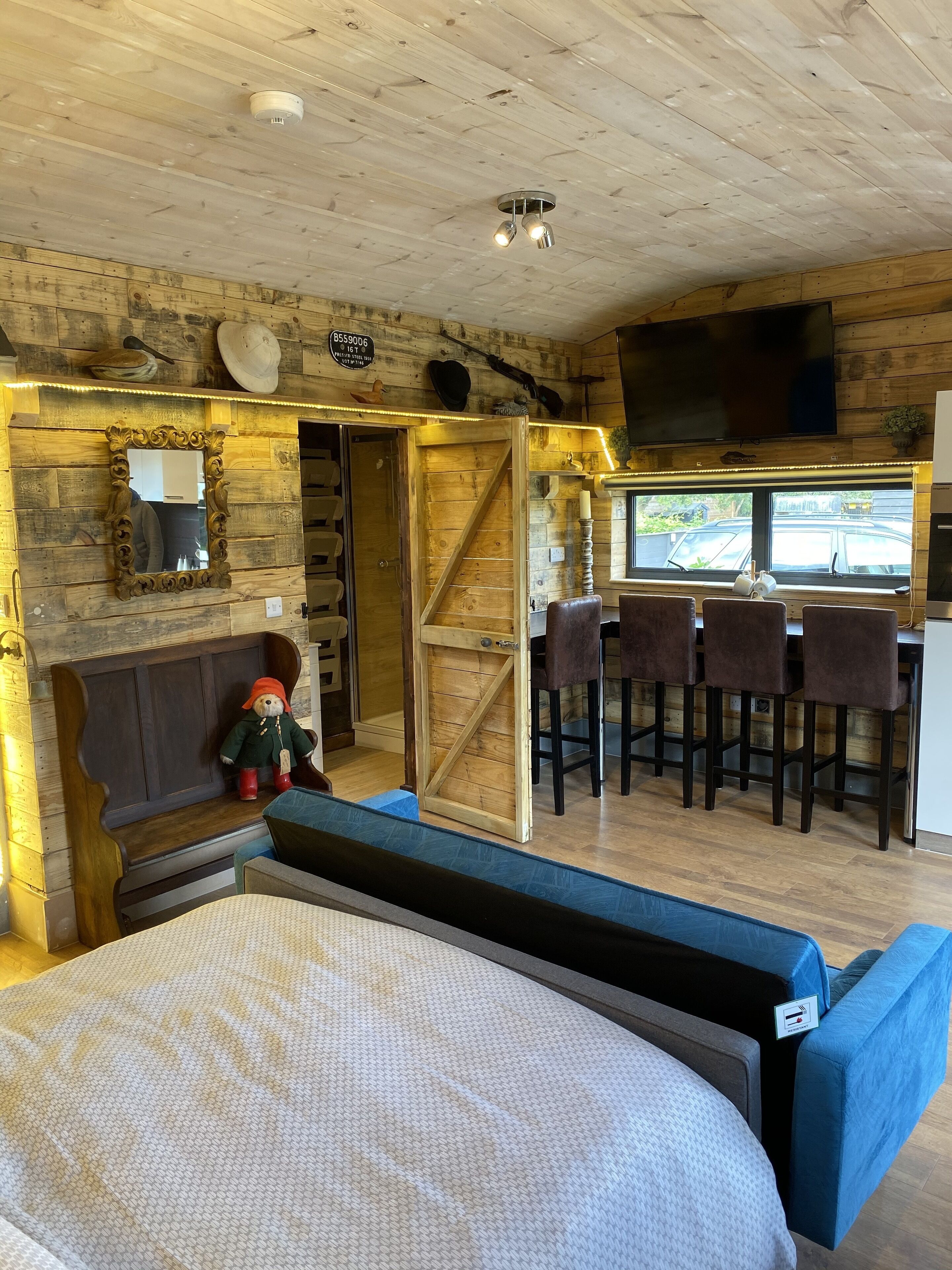 A cozy room with a wooden ceiling, a large bed, a small table with a teddy bear, and a bar area with chairs and a TV.