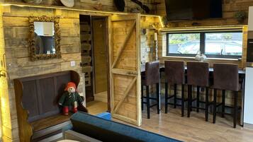 A cozy room with a wooden ceiling, a large bed, a small table with a teddy bear, and a bar area with chairs and a TV.