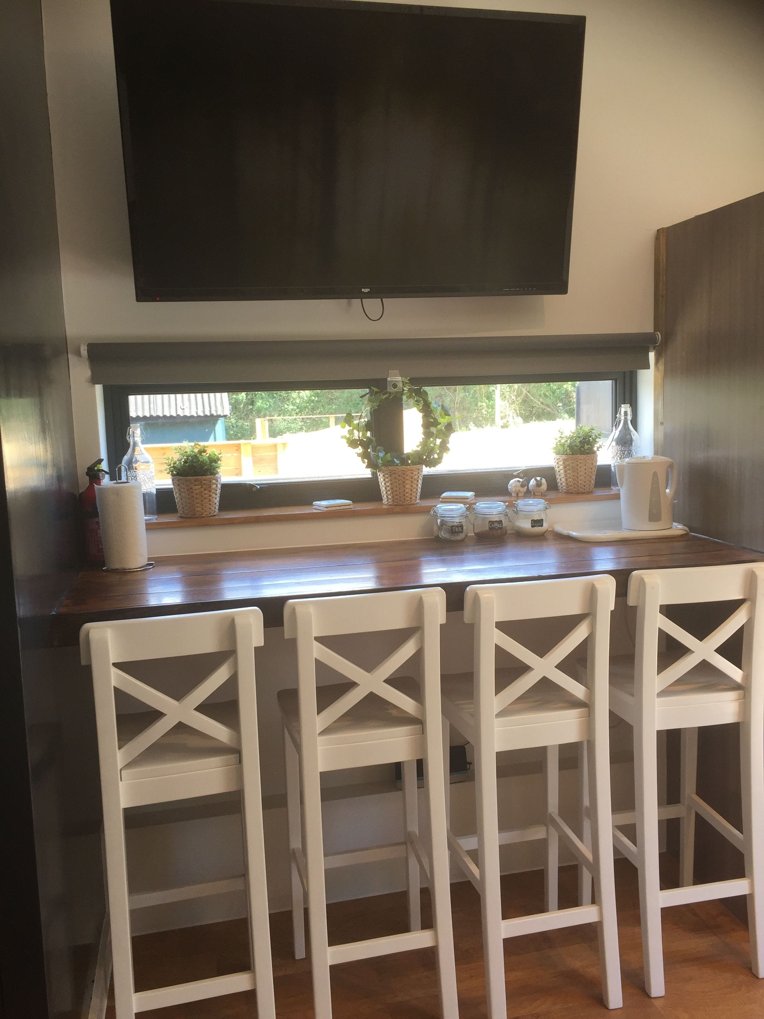 A kitchen with a large TV mounted above a window, a wooden countertop, and four white chairs with a cross-back design.