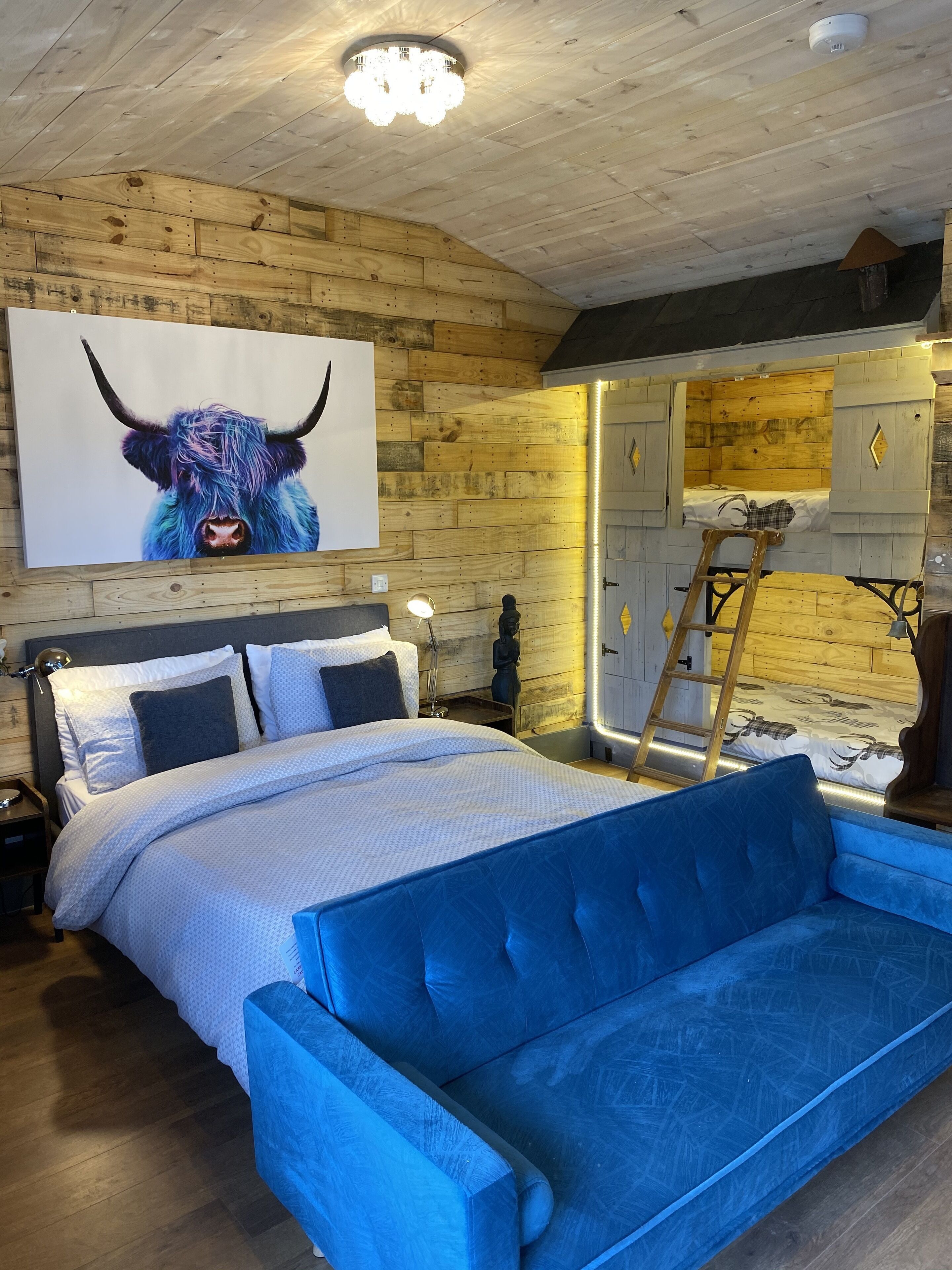 Un dormitorio acogedor con techo de madera, una cama con cabecera tapizada en azul, una mesita de noche, una lámpara y una obra de arte en la pared con motivo de un toro.