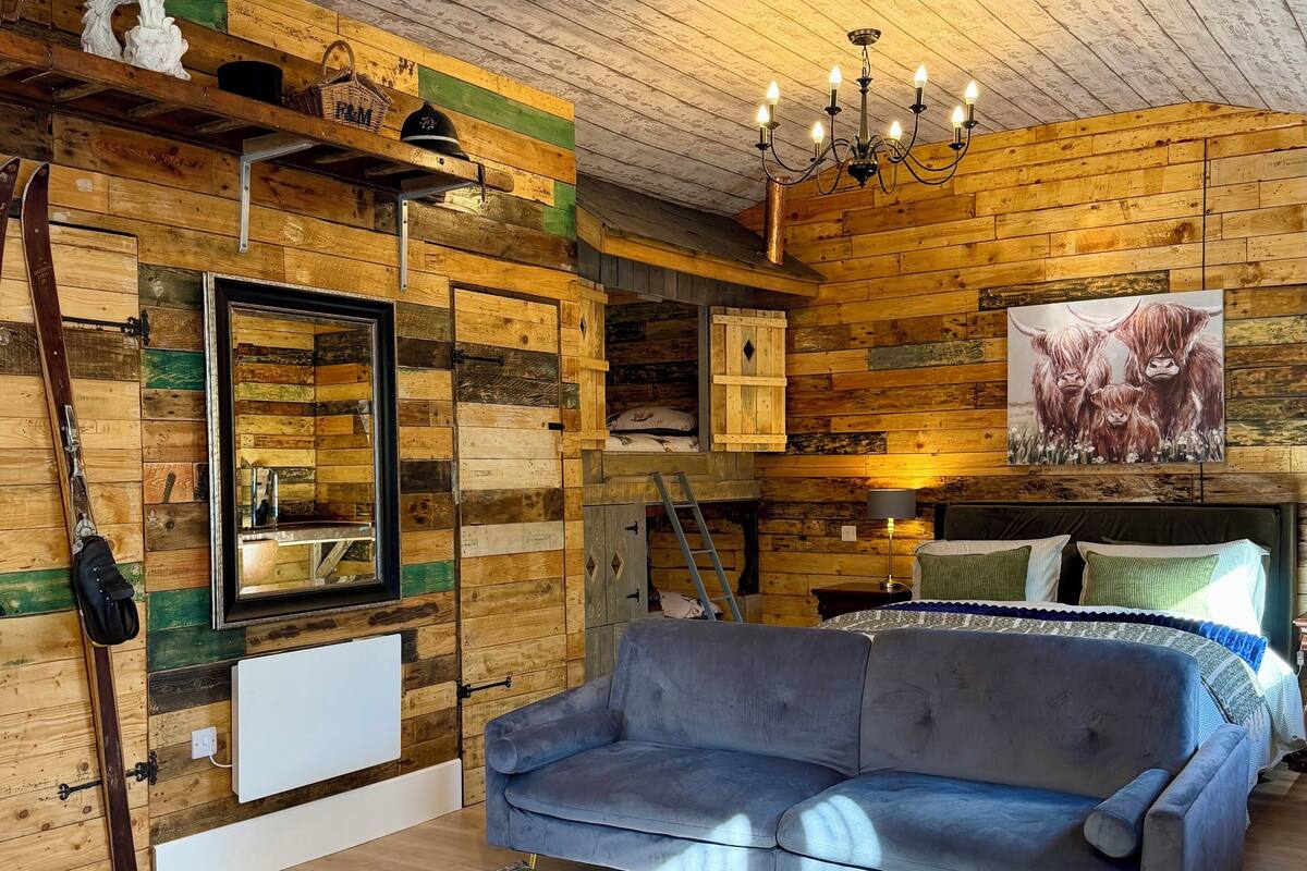 L’intérieur d’un chalet rustique, avec un canapé bleu, une malle verte, des murs en bois, un lustre et un tableau représentant des yaks.