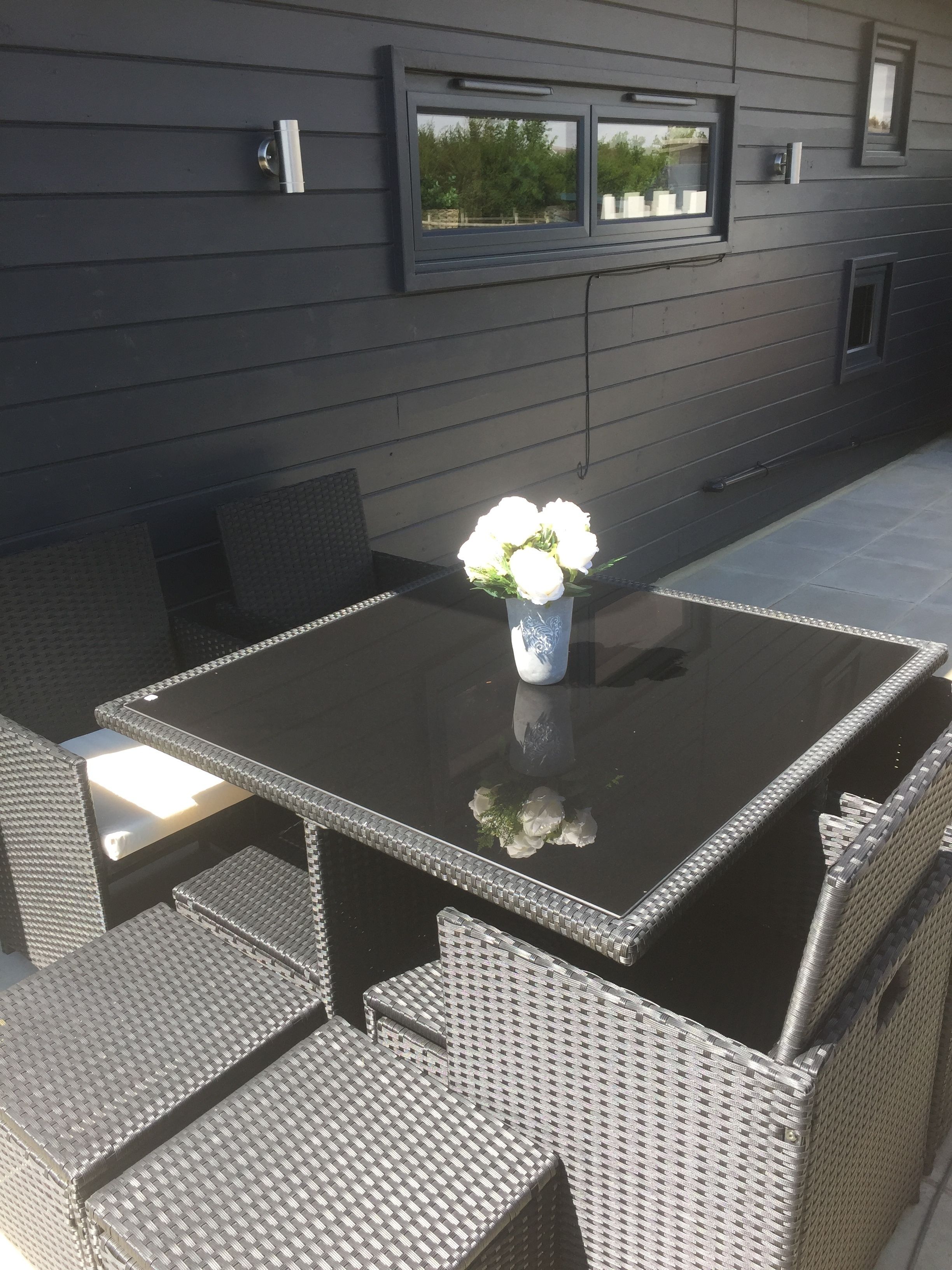 Een terras buiten met een zwarte tafel en rieten stoelen, een witte vaas met bloemen, en een donkere muur met ramen en een deur.