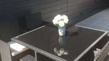 Een terras buiten met een zwarte tafel en rieten stoelen, een witte vaas met bloemen, en een donkere muur met ramen en een deur.