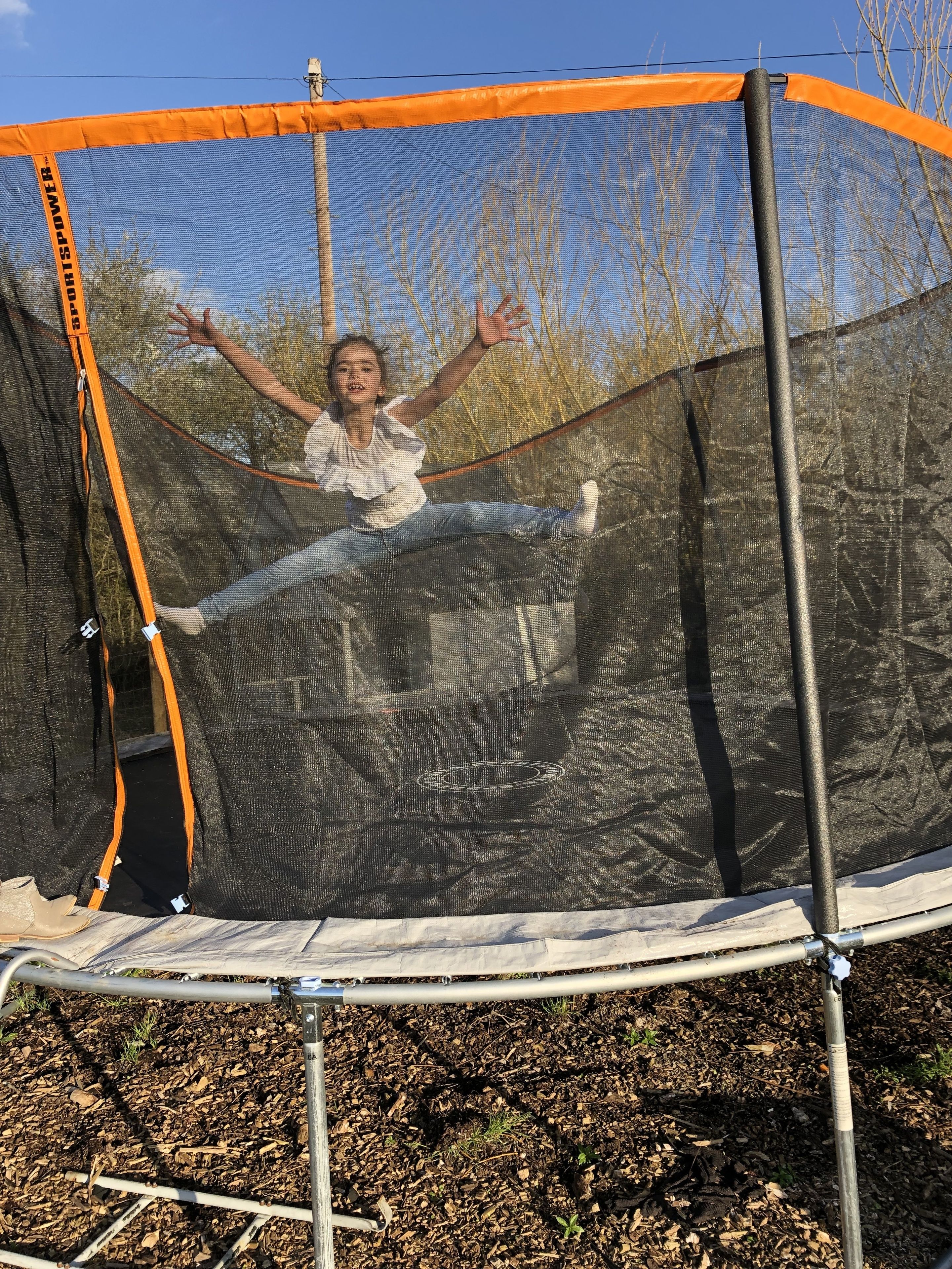 A trampoline with an orange safety net, set up outdoors with a metal frame and surrounded by mulch.