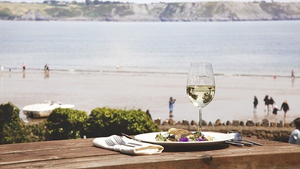 A wooden table with a plate of food, a glass of white wine, and cutlery, overlooking a beach with people and a boat in the distance. - Oxwich Bay Hotel (Swansea)