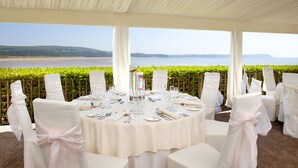 A round table set for a formal event with chairs covered in white fabric and pink sashes, overlooking a beach and sea. - Oxwich Bay Hotel (Swansea)