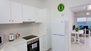 Cocina moderna con armarios blancos, estufa, horno y refrigerador. Hay un comedor con mesa y sillas, y vistas al mar a través de amplios ventanales.