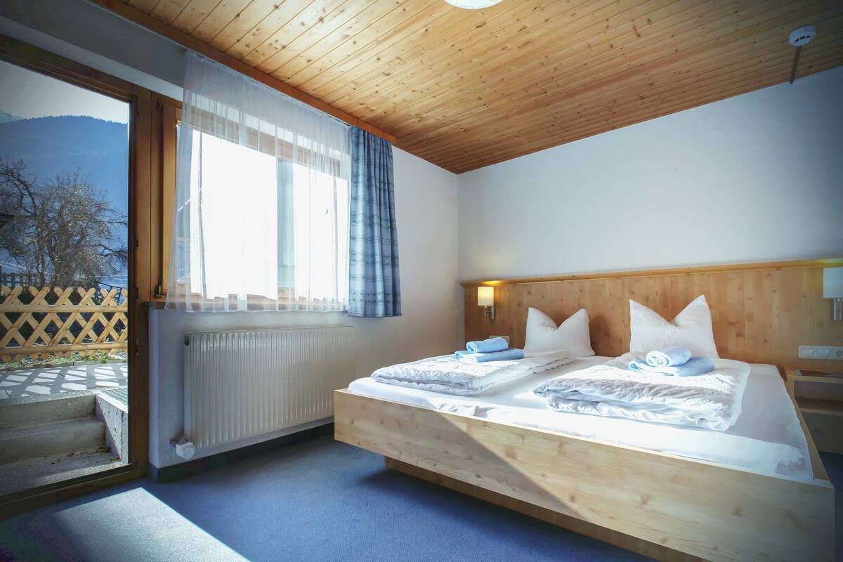 Un dormitorio con techo de madera, un ventanal con cortinas, una cama con ropa de cama blanca y vistas a montañas.