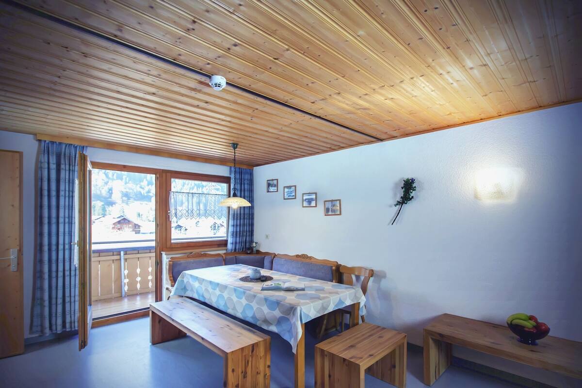 Una habitación con techo de madera, una mesa de comedor con bancos, una ventana con cortinas y una lámpara fijada a la pared.
