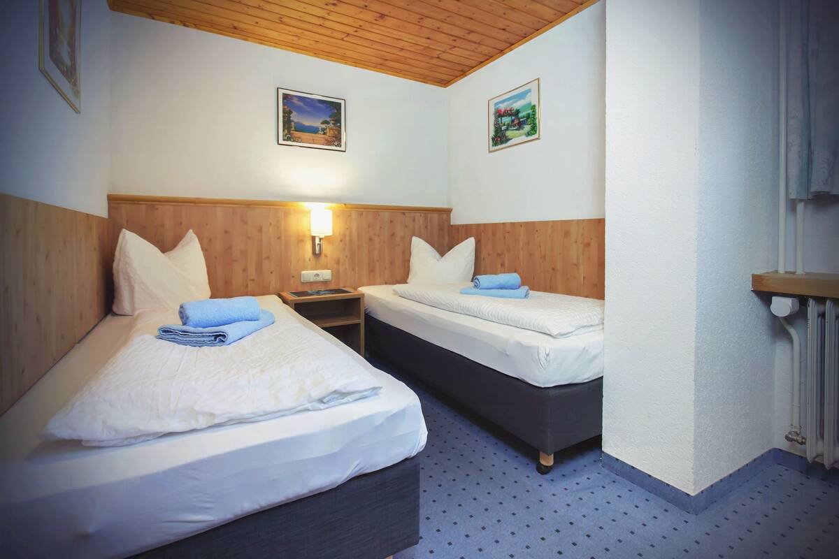 Habitación de hotel con dos camas, cabeceros de madera, una mesita de noche, una l ámpara y dos cuadros en la pared.