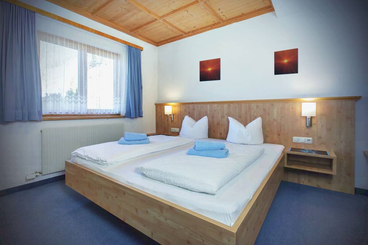 Cama doble con sábanas blancas y toallas azules, cabecera de madera, dos lámparas de pared y una ventana con cortinas.
