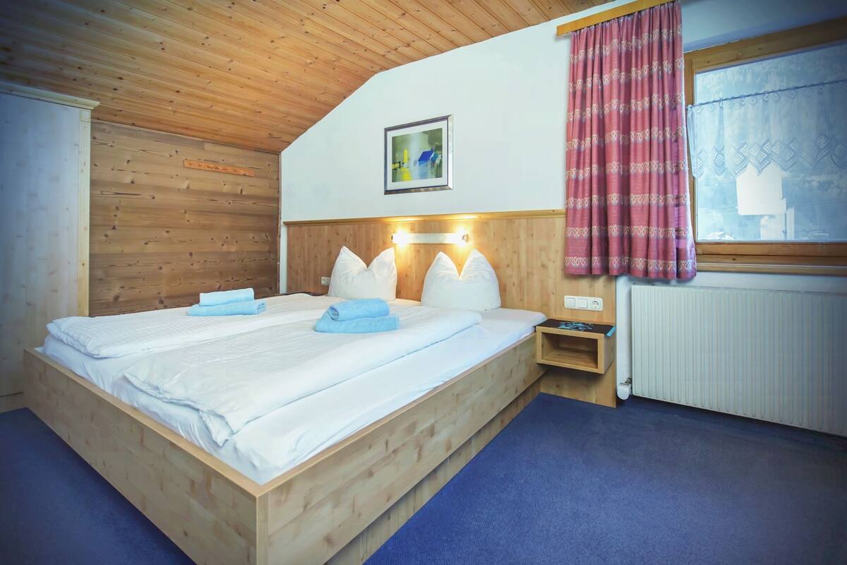 Una cama de madera con sábanas blancas, una mesita de noche, una lámpara fijada a la pared y una ventana con cortina estampada.