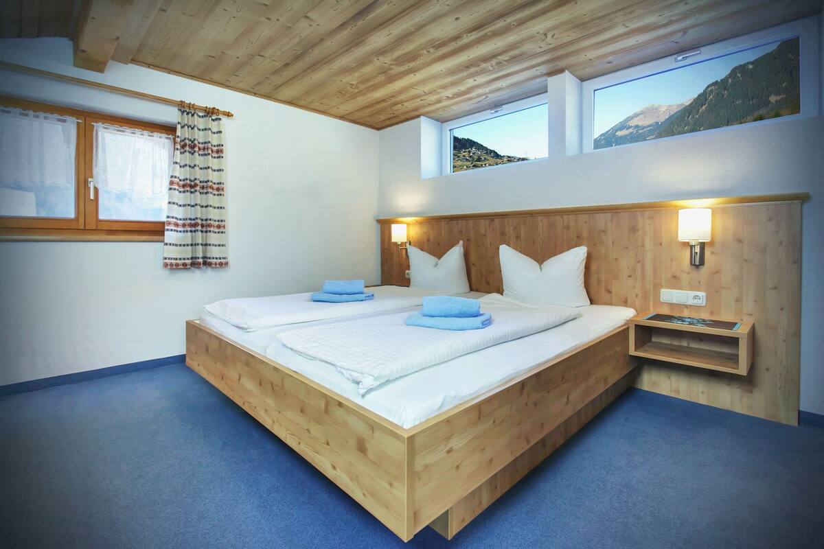 Cama doble con cabecera y piecera de madera, dos mesitas de noche, una ventana con cortinas y vistas a la montaña.