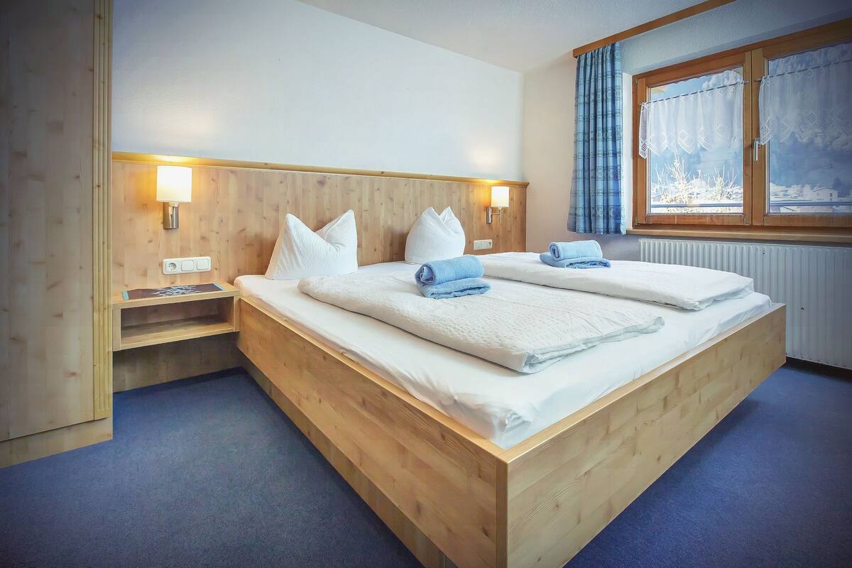 Una cama de madera con dos almohadas, una mesita de noche, una lámpara y una ventana con cortinas.