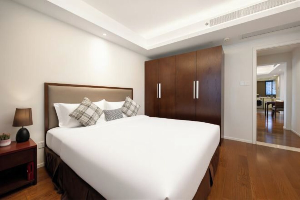 A hotel room with a large bed, wooden wardrobe, bedside table with lamp, and a view into the kitchen area.
