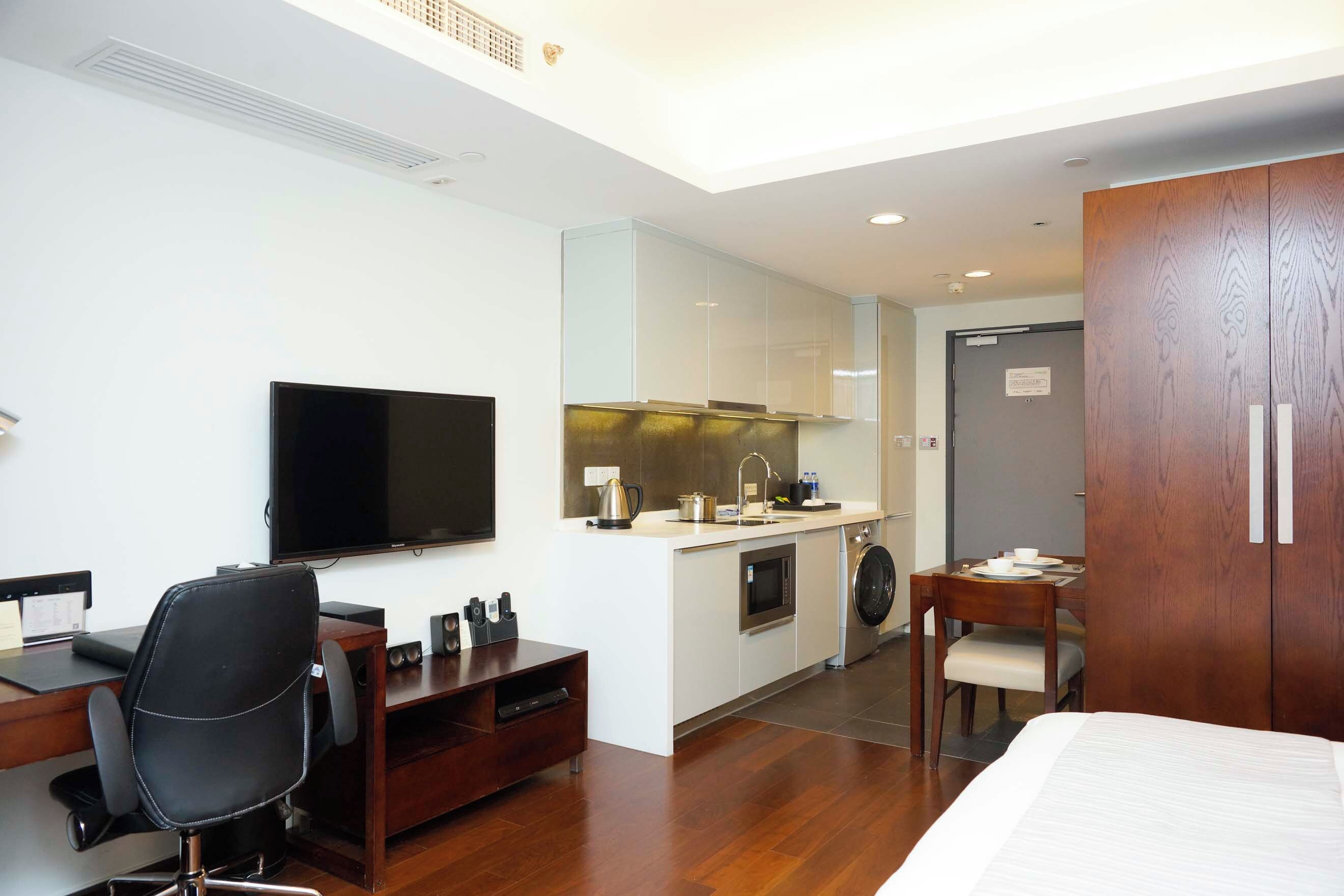 A compact hotel room with a bed, a desk with a chair, a television, a kitchenette, and a wardrobe.