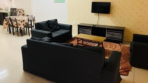 Een woonkamer met een zwarte bank, een klein bijzettafeltje, een aan de muur bevestigde televisie en een vloerkleed met een patroon.