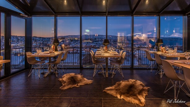 A restaurant with a panoramic city view, modern furniture, and fur rugs on the floor. - Breidablikk Gjestehus (Narvik)