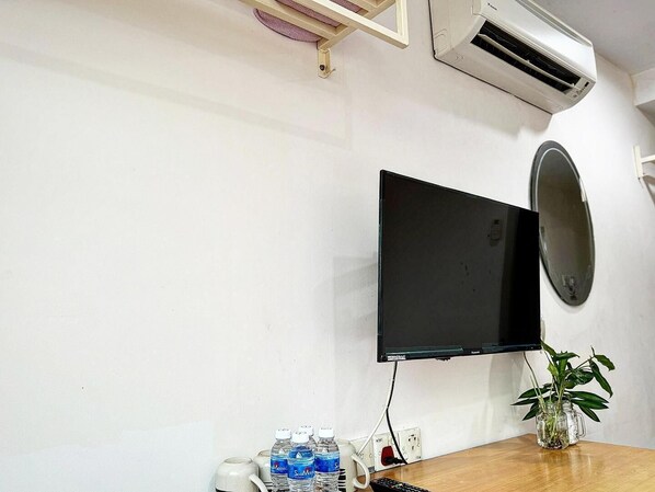 A room with a mounted flat-screen TV, a wooden table with a mug, water bottles, a remote control, and a potted plant. - Town Inn Hotel (Jerantut)