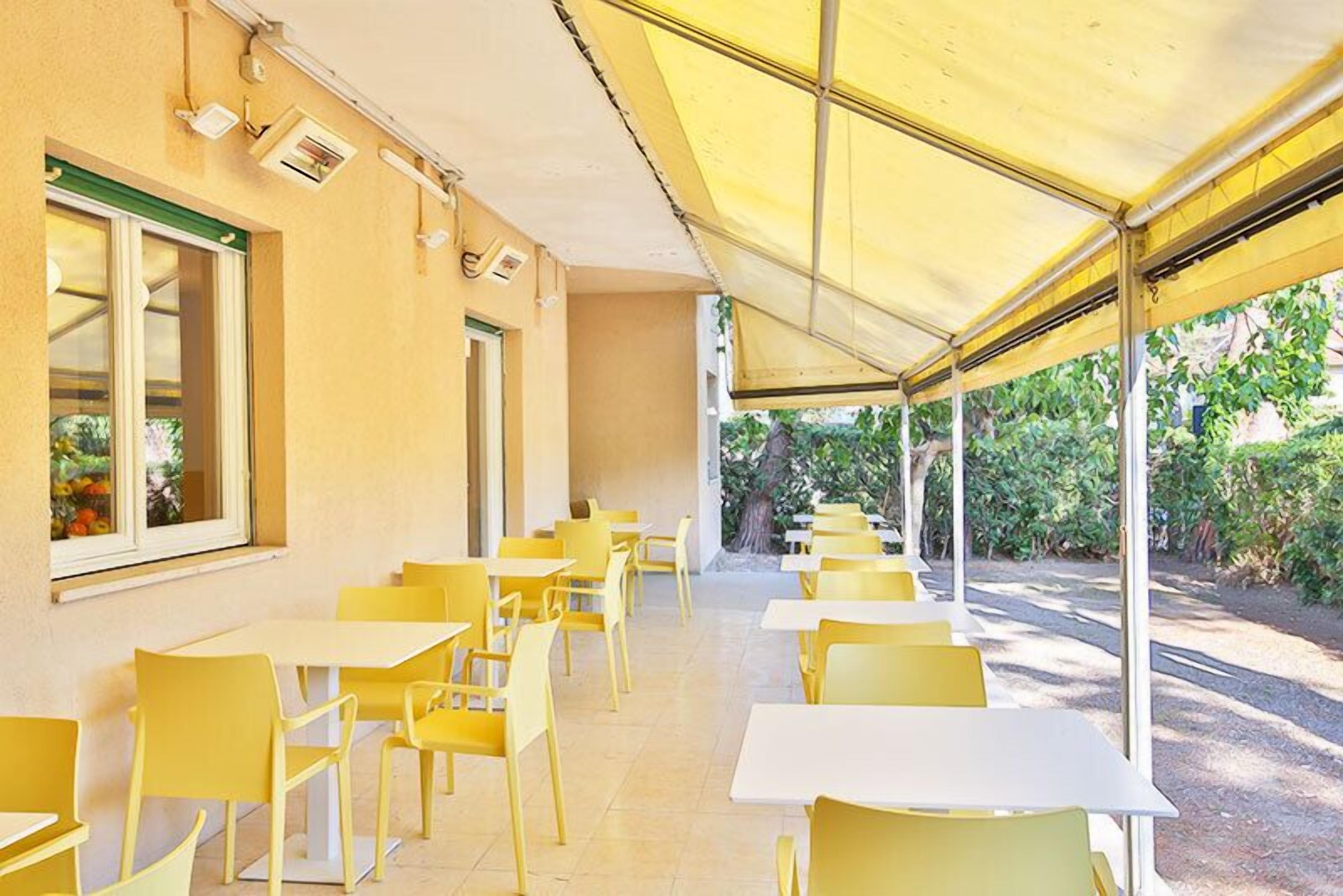 Un café al aire libre con mesas y sillas amarillas, cubierto por un toldo amarillo y rodeado de follaje.