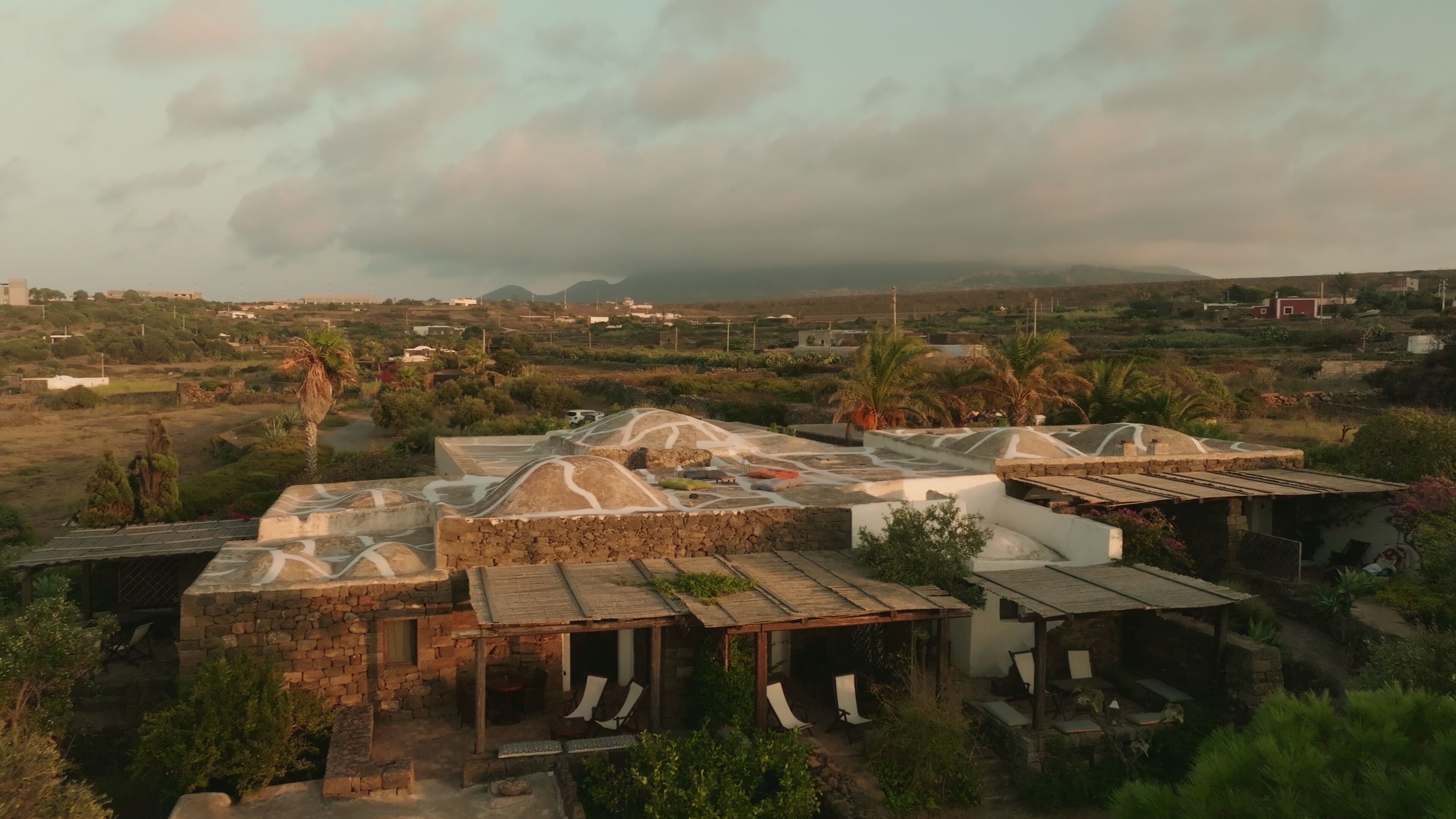 A rustic village with stone buildings and thatched-roof huts, surrounded by greenery and distant mountains.