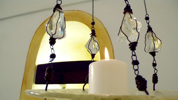 A candle on a white stand with a clear glass bowl, a white candle, and a window with an arched frame.