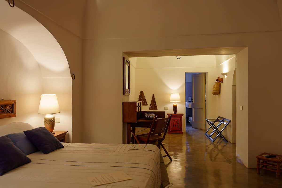 A hotel room with a large bed, two bedside lamps, a wooden headboard, a desk with a chair, and a small stool.