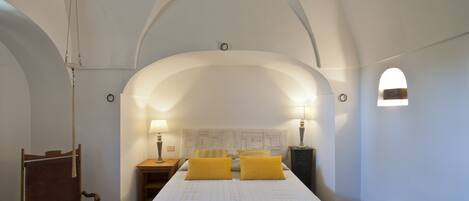 A room with a large bed, two yellow pillows, a wooden chair, a nightstand, and a wall-mounted lamp.
