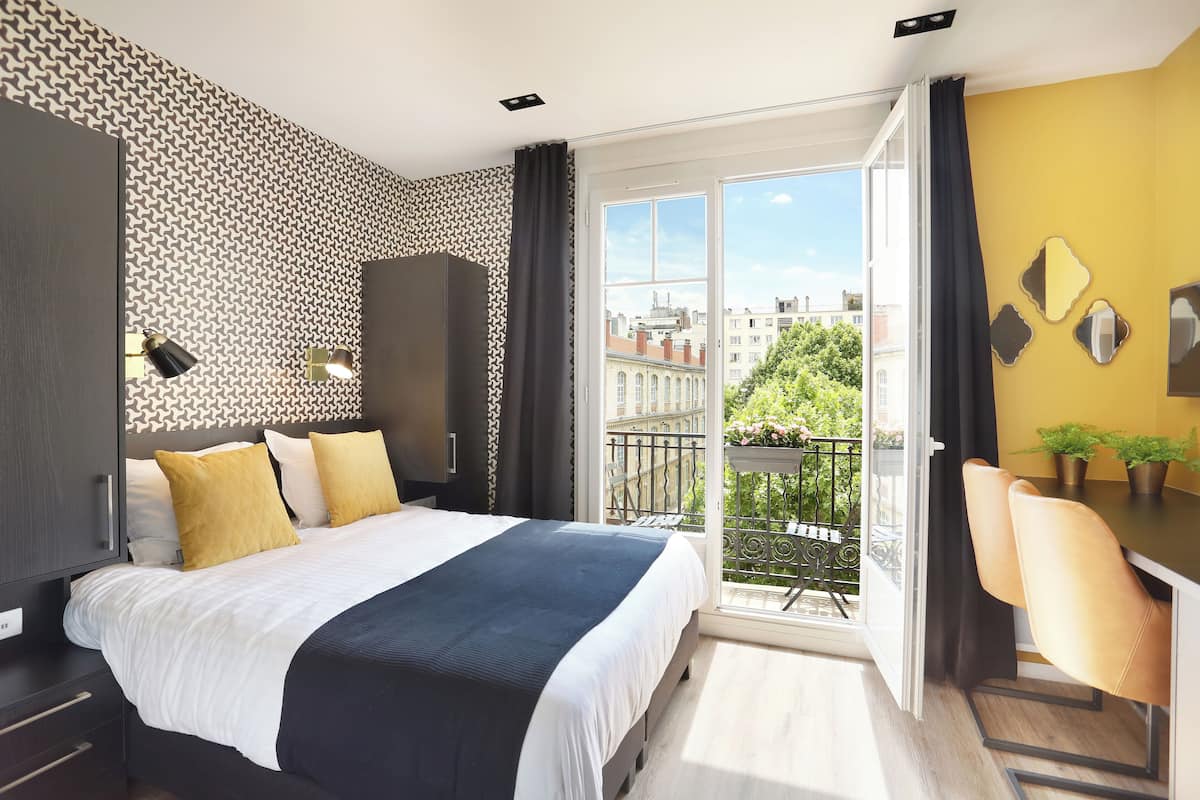 A modern hotel room with a large bed, a desk, and a balcony with a city view.