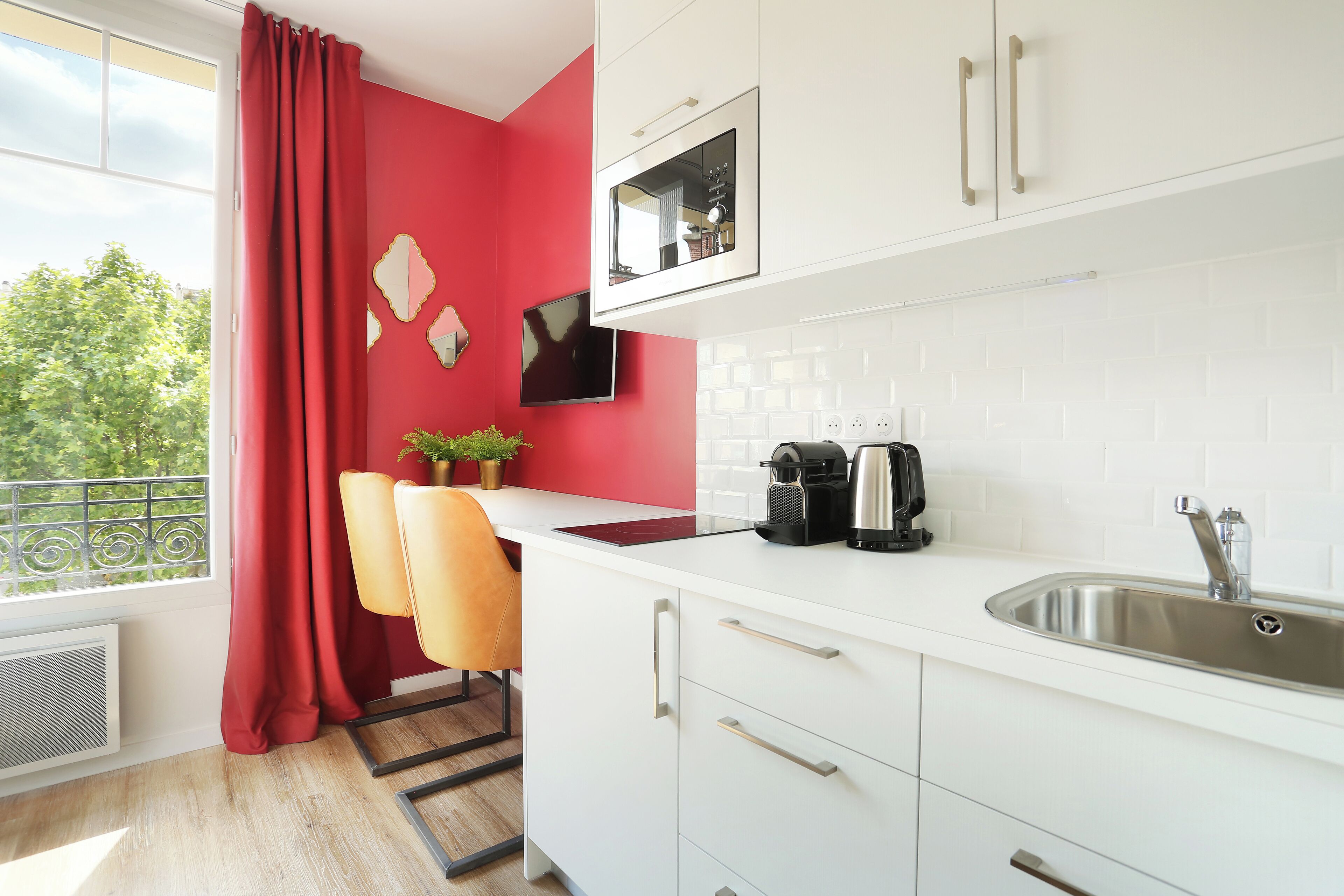 A modern kitchen with white cabinets, a red accent wall, a window with red curtains, and a small potted plant on the counter.