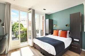 Un dormitorio con una cama grande, un armario, un escritorio y un balcón con vistas a edificios y árboles.