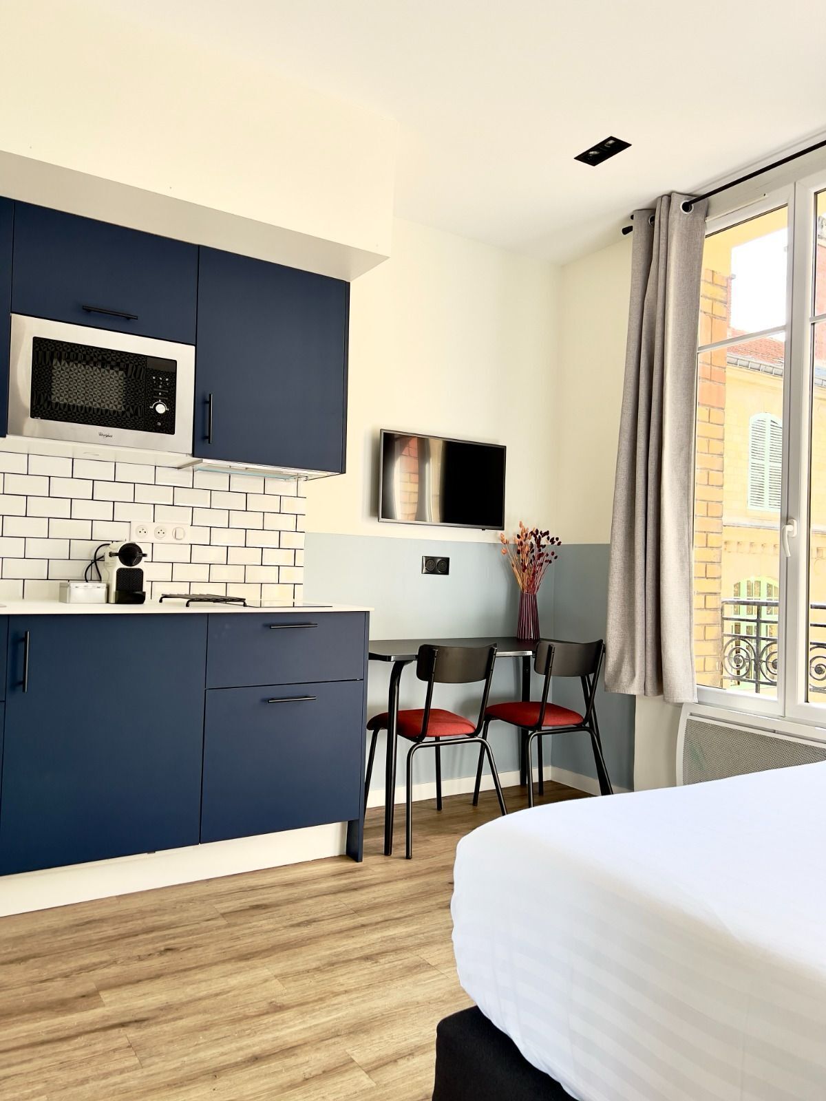 A compact hotel room with a kitchenette, a bed, a dining area, and a window with curtains.