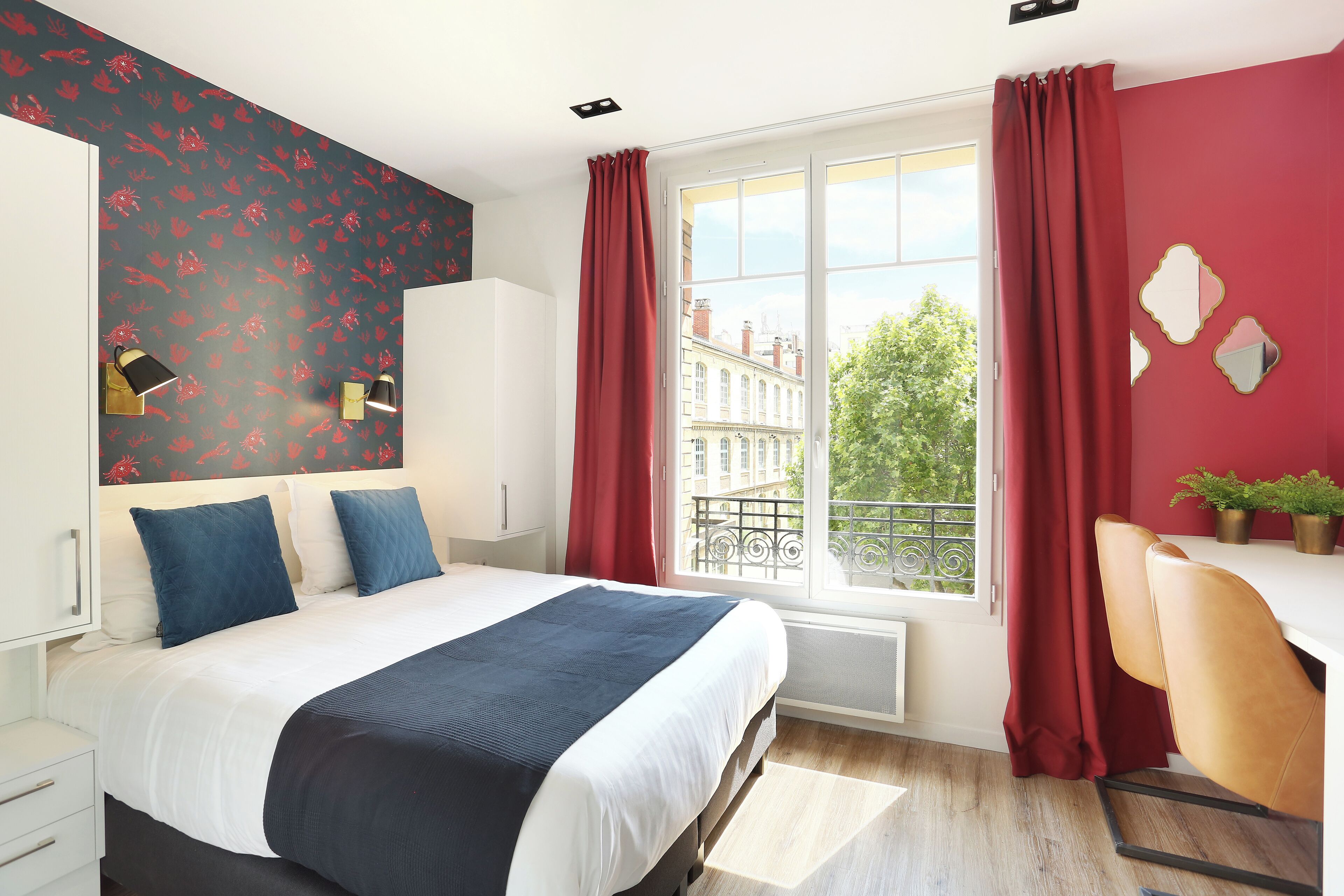 Camera da letto moderna con un letto grande, una scrivania e vista sulla città attraverso una finestra con tende rosse.