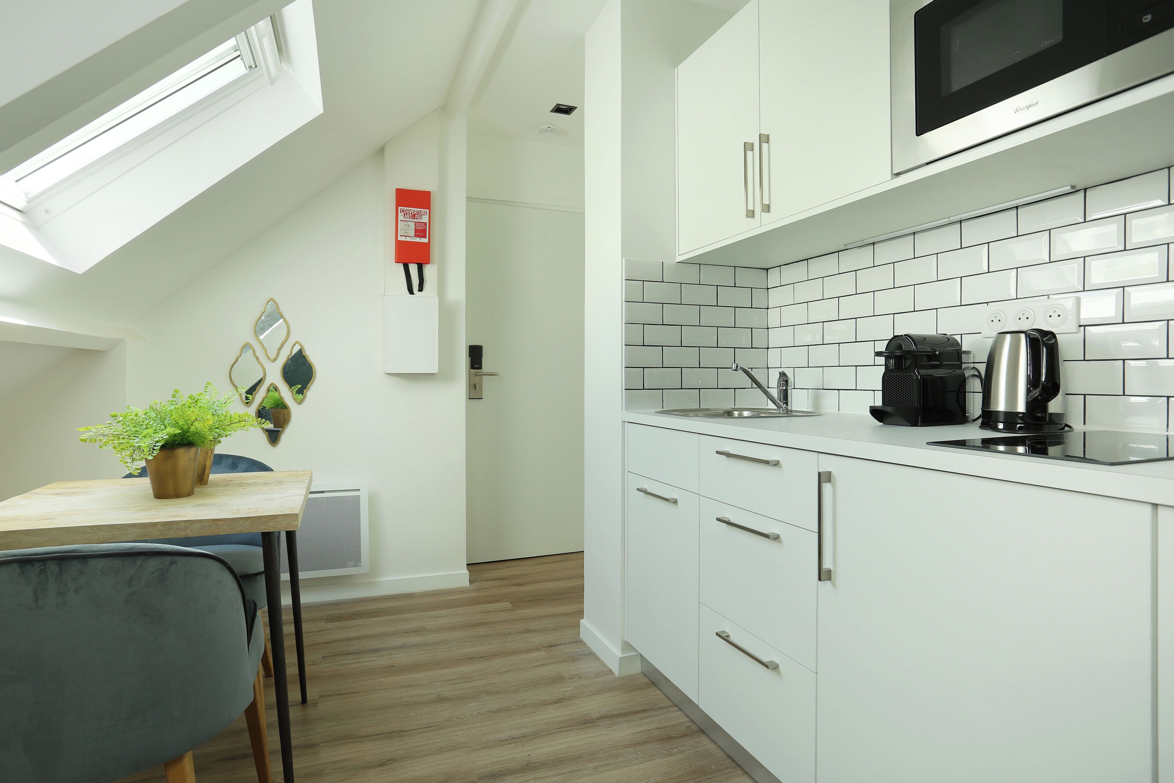 A modern kitchen with white cabinets, a microwave, and a coffee maker. There is a small table with a plant and a chair.
