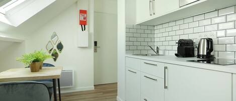A modern kitchen with white cabinets, a microwave, and a coffee maker. There is a small table with a plant and a chair.