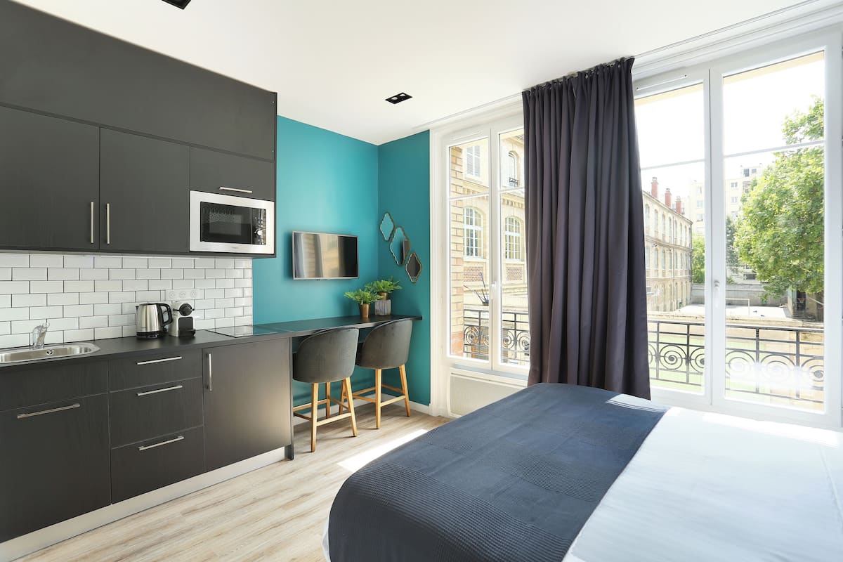 A modern kitchen with dark cabinets, a microwave, and a sink. There is a bed with a large window and a view of buildings outside.