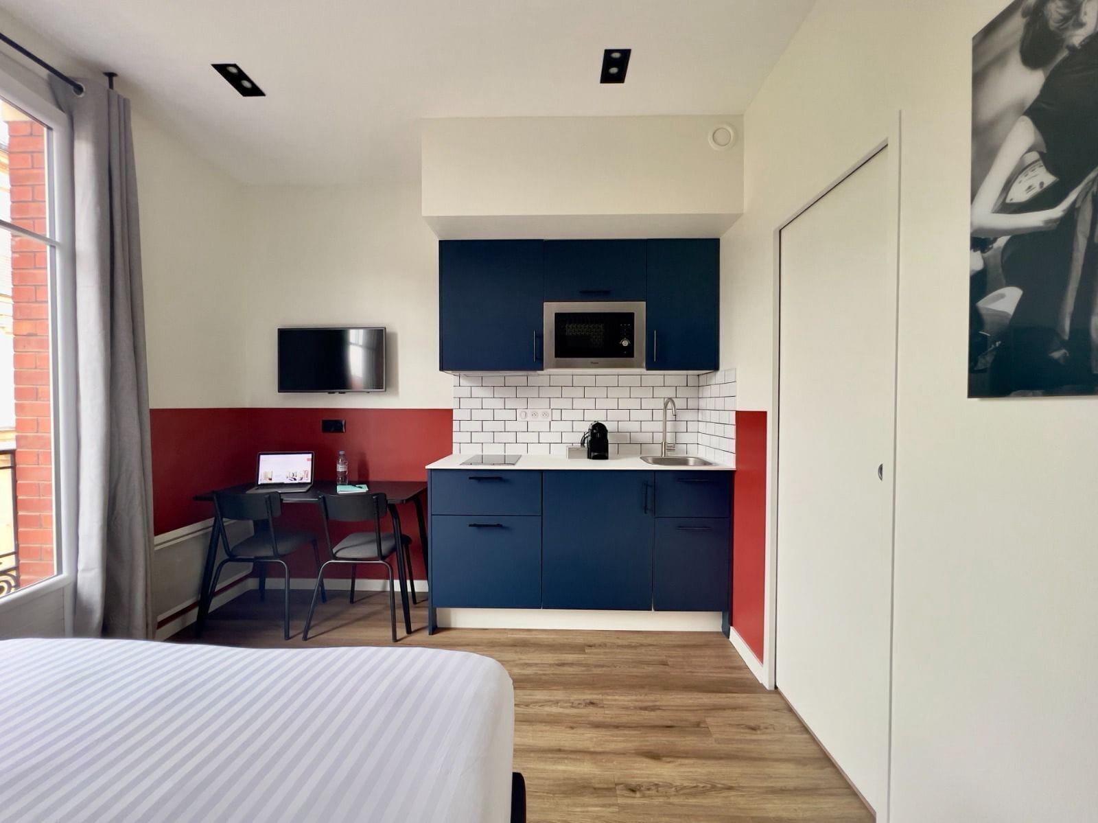 A compact hotel room with a bed, a desk, a kitchenette, and a TV.