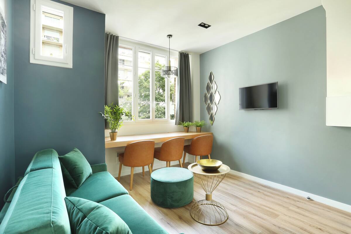 A modern living room with a teal sofa, a dining area with a table and chairs, a TV mounted on the wall, and a window with curtains.